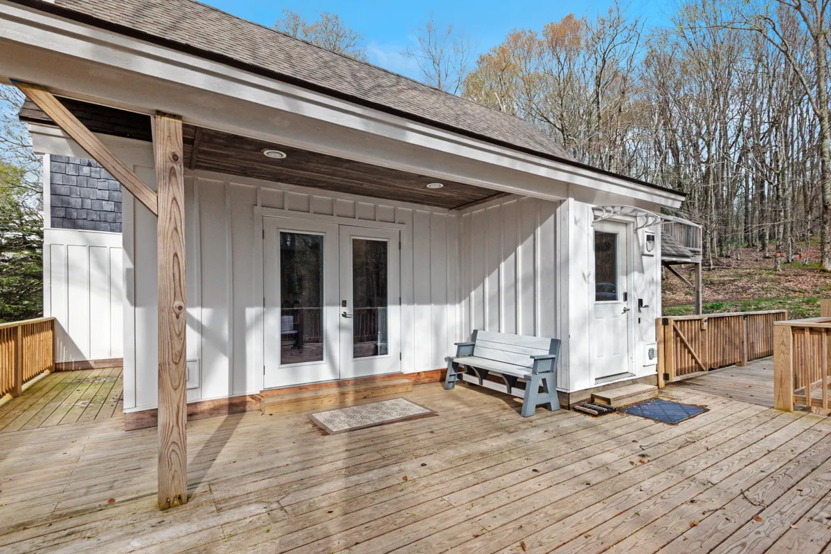 The image shows a serene wooden deck with double doors opening to the house, a bench, and mature trees surrounding the space. The setting is tranquil, inviting, and perfectly suited to morning coffee or evening gatherings amidst nature.