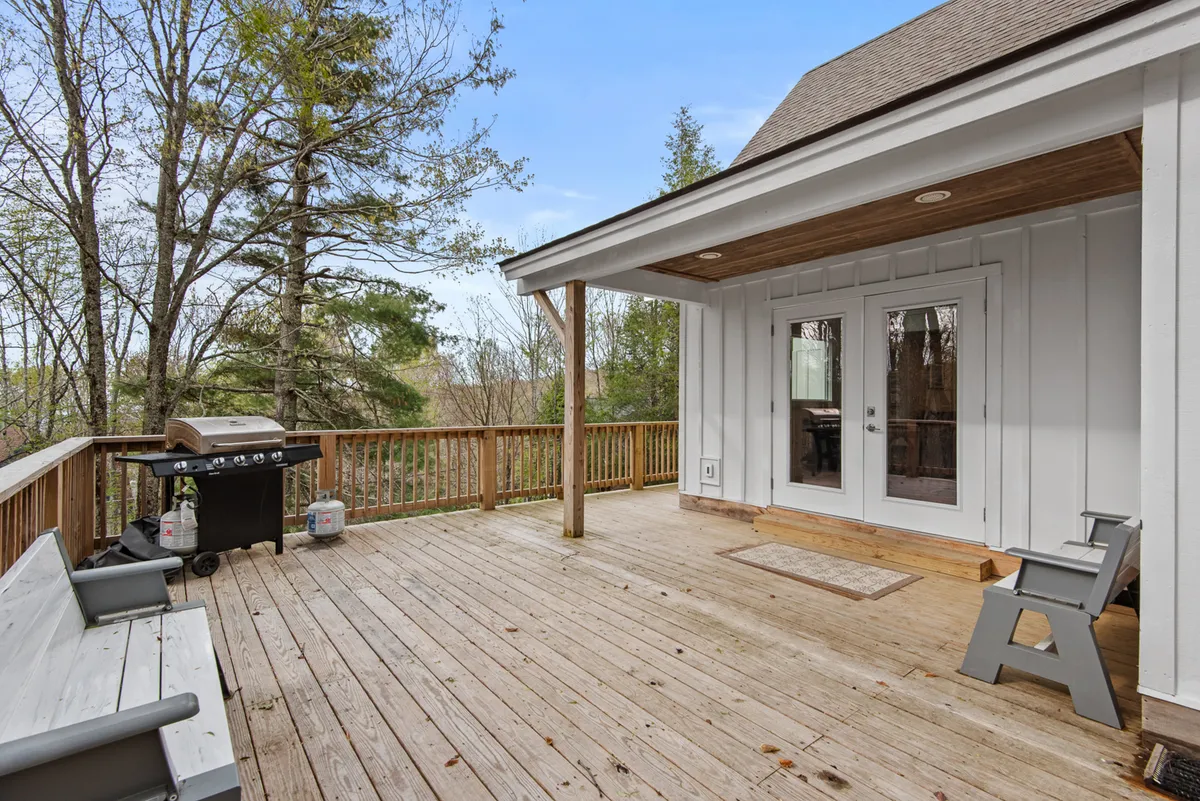 A spacious wooden deck with a grill and sturdy wooden railing, double doors opening from the main living area, and ample space for socializing and outdoor dining. The setting is surrounded by trees, capturing a relaxed mountain vibe perfect for gatherings.