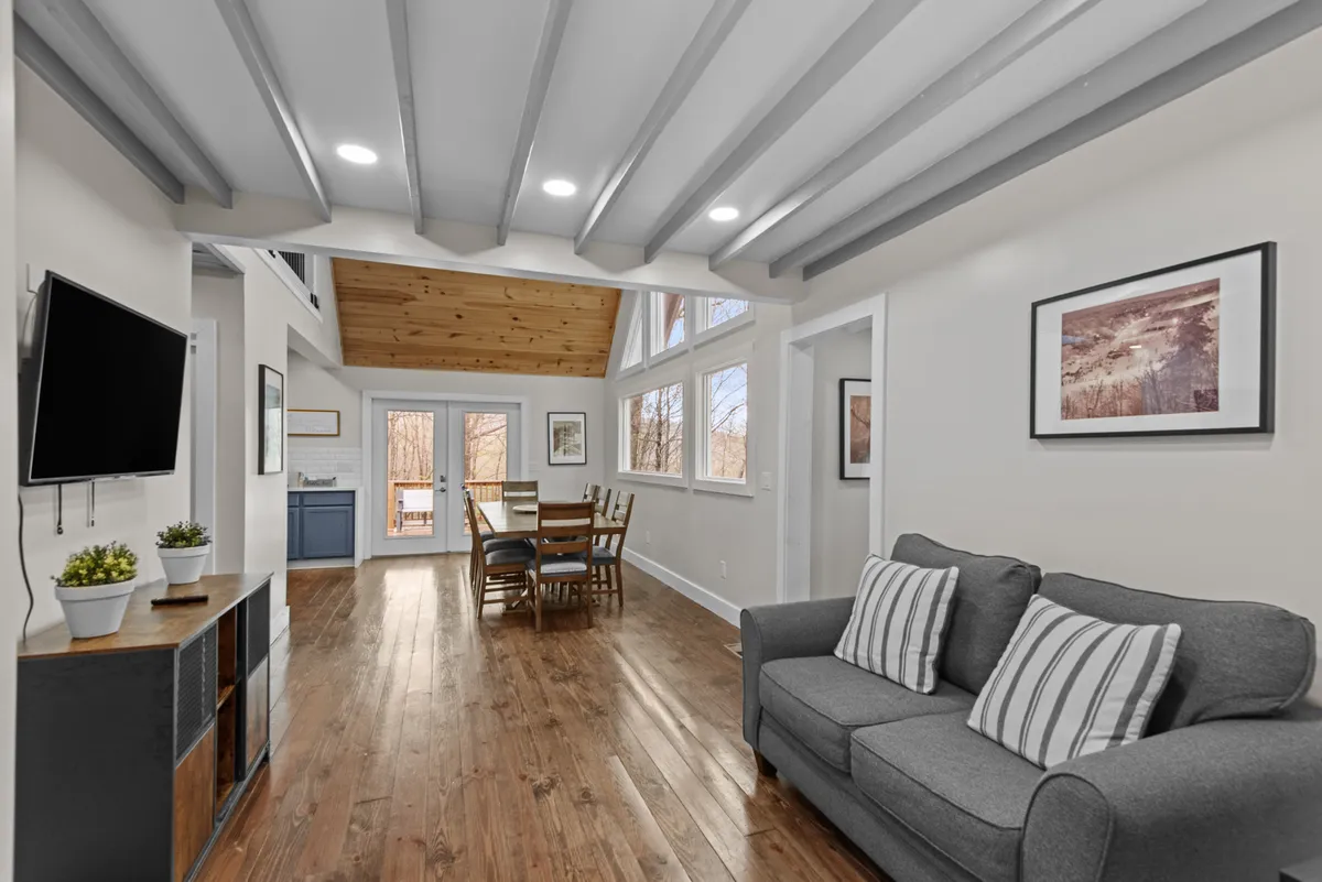 An inviting open-plan living and dining space, complete with a comfortable sofa, striped cushions, a family-sized dining table, large windows streaming in natural light, and exposed wooden beams overhead. The room exudes warmth, ideal for cozy gatherings or relaxed evenings.