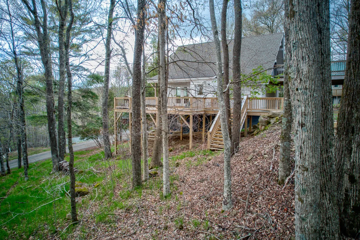 A broad wooden deck extends from the house, surrounded by lush trees and offering panoramic views. The space is set up for gatherings and relaxation, with hints of both sunrise and sunset experiences.