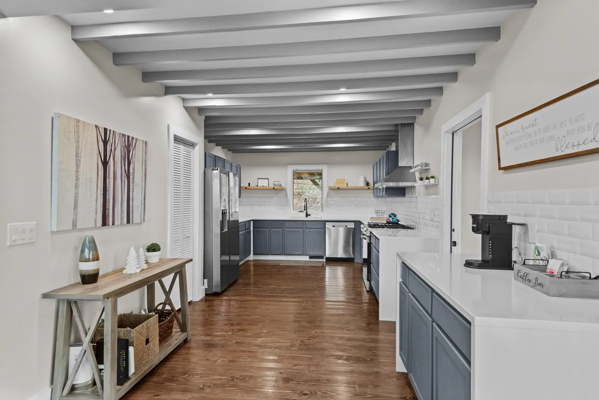 This image showcases a stylish, well-lit kitchen with a central island, stainless steel appliances, and wooden beams overhead. Artwork on the wall lends a welcoming, homely touch. The space is clearly designed for both form and function, inviting for cooking and gathering.