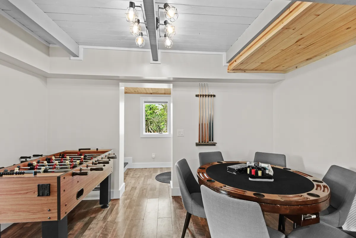 A vibrant basement game room featuring a foosball table, a poker table, pool table cues, and a large window letting in natural light. The space is arranged to invite friendly competition and social gatherings, with ample room for guests to participate in games or relax nearby.