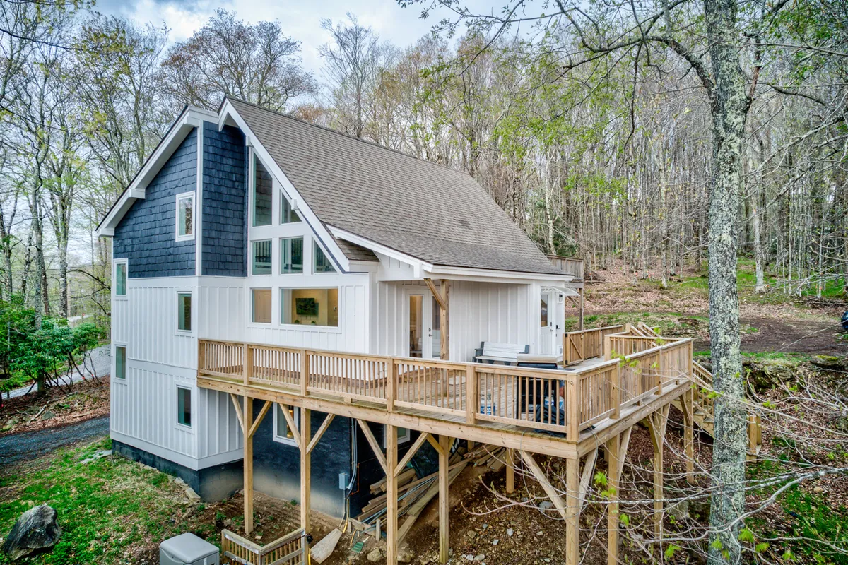 The image showcases the exterior of a beautifully renovated mountain home nestled in lush natural surroundings. A spacious wooden deck invites guests to relax, with large windows offering glimpses of the inviting interior. The setting is tranquil, highlighted by sunset-painted skies and the promise of stargazing at night.