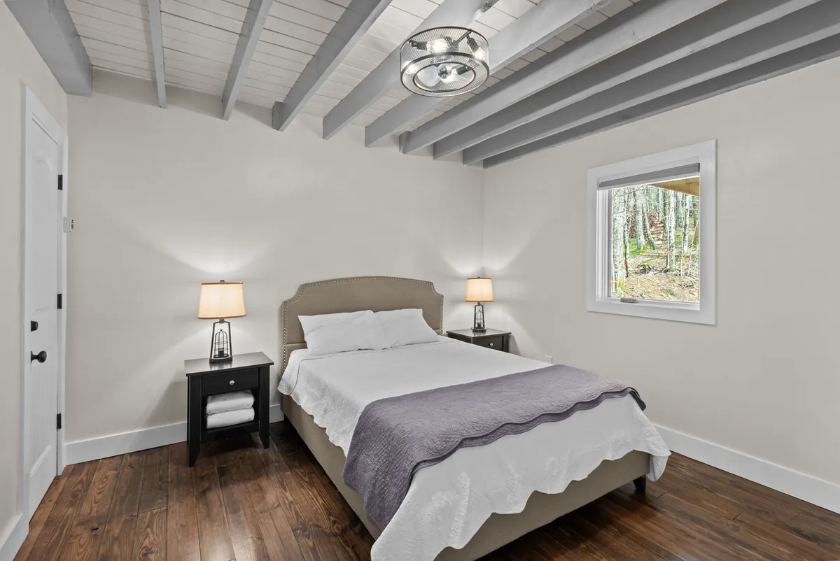 A tranquil bedroom with a bed dressed in soft linens, flanked by nightstands with lamps. Sunlight pours in through a window revealing lush greenery outside, emphasizing a restful and inviting atmosphere.
