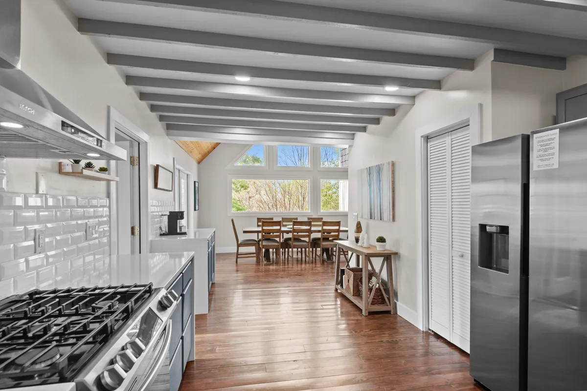 A wide-angle view merging the kitchen island, dining table, large windows, and wooden beams on the ceiling. This image beautifully demonstrates the open-concept layout, where kitchen, dining, and living spaces flow together seamlessly, awash in natural light.