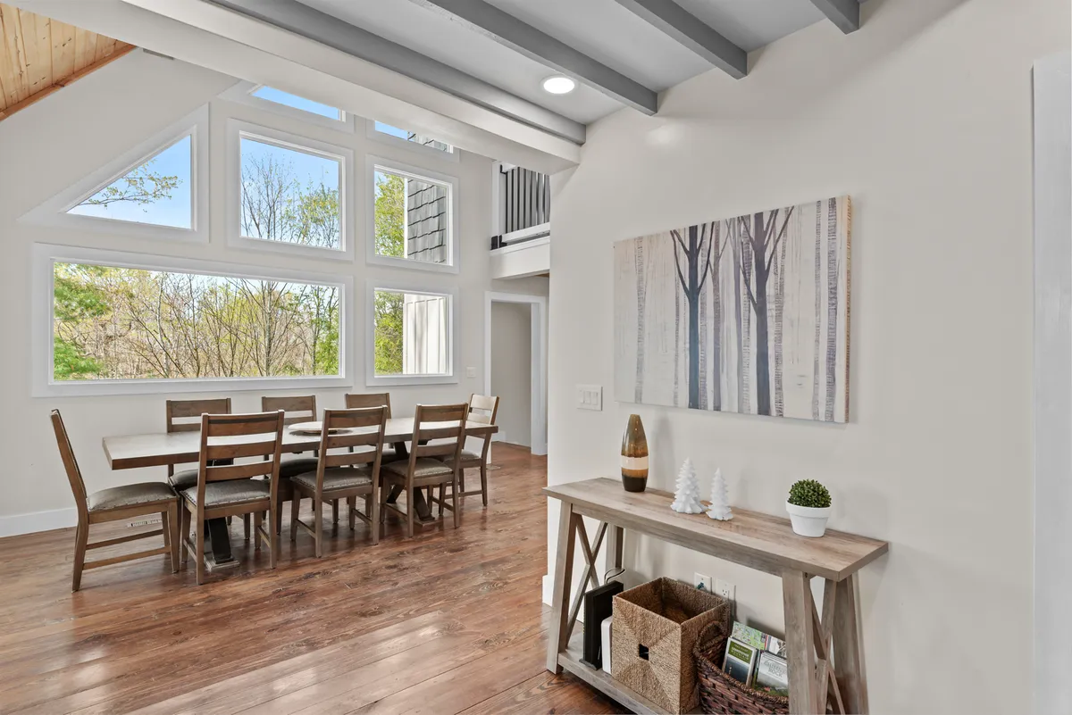 A spacious dining area featuring a large table set beneath expansive windows, allowing an abundance of natural light to fill the room. Artwork adorns the wall, and the setup is ideal for group gatherings, whether for meals, conversations, or celebrations.