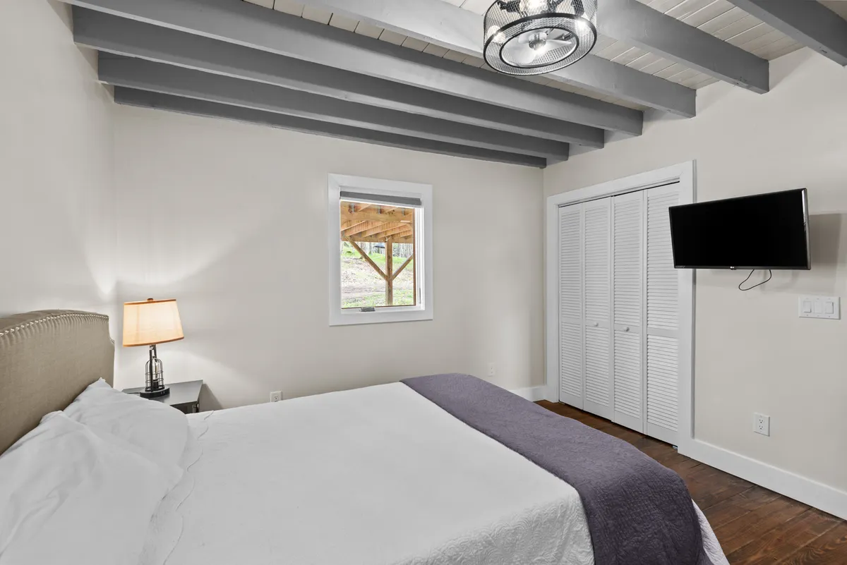 A comfortable bedroom featuring white linens with a purple throw, a window looking out onto nature, and a bedside lamp. The layout is conducive to both relaxation and leisure, offering an inviting space for unwinding at any time of day.