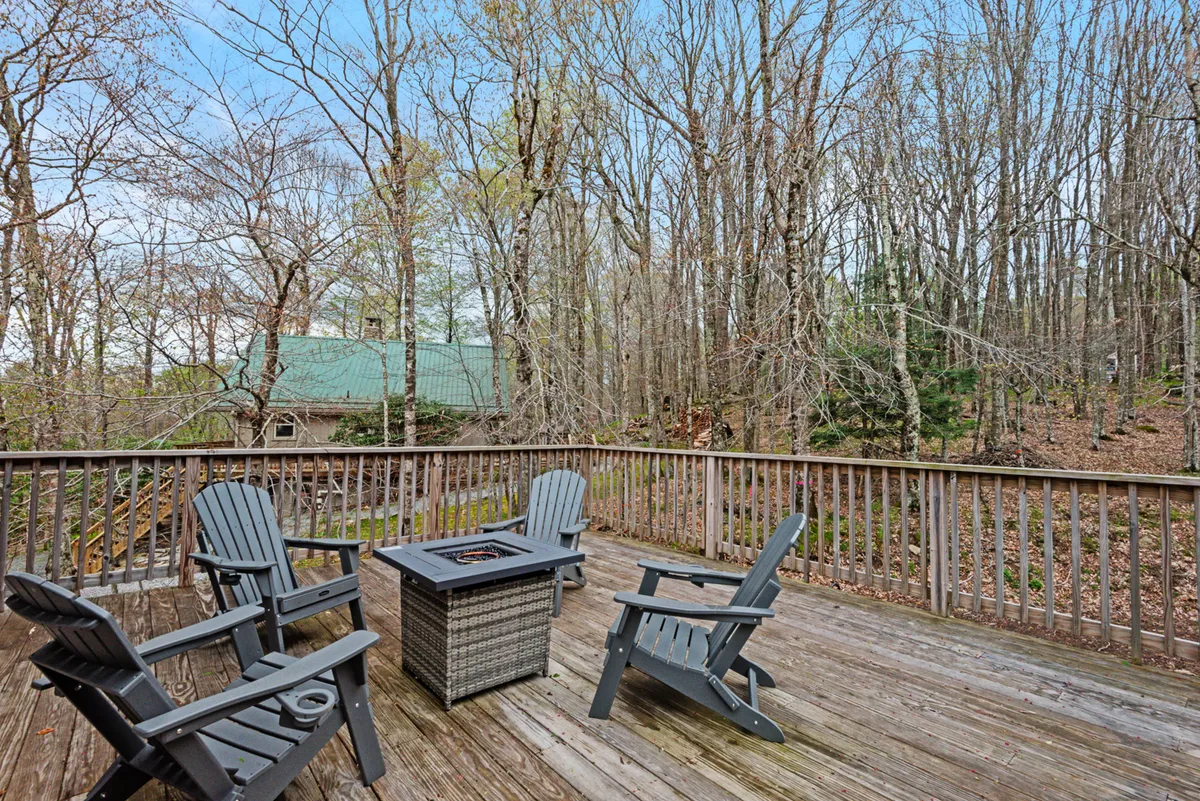 A spacious wooden deck features a central fire pit surrounded by comfortable outdoor seating, with lush trees and forest in the background. The setting is ideal for evening gatherings, relaxation, and enjoying nature after a day of adventure.