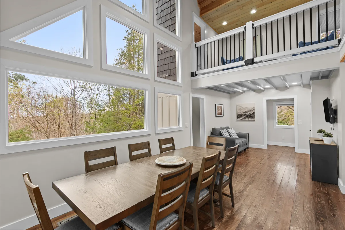 A warm dining area is highlighted here, centered around a generous table with seating for a group. Large windows surround the space, bringing in natural light and framing scenic views; a lofted area above adds architectural interest, and the mood is perfect for memorable family celebrations or meals.